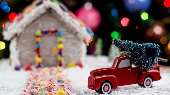 Ein rotes Spielzeugauto hat einen Weihnachtsbaum geladen und steht in der Vorfahrt eines Lebkuchenhauses.