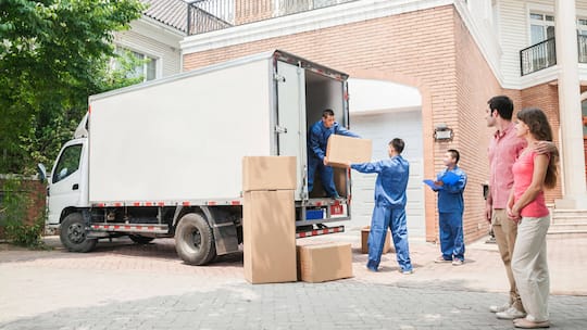 Junges Ehepaar zieht in das geschenkte Haus ein und steht vor dem Umzugswagen