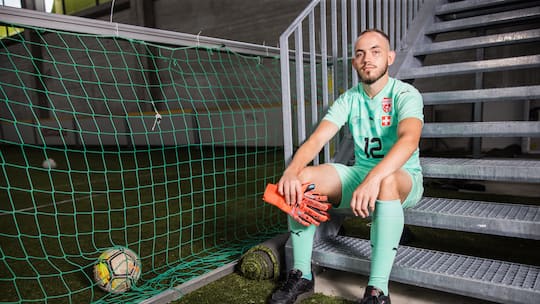 Patrick Allenbach sitzt im Fussballtenue auf einer Treppe in einer Sporthalle, offener Blick in die Kamera, Goalie-Handschuhe in der Hand