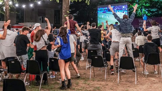 Fussballfans feiern ausgelassen ihre Mannschaft bei einem Public Viewing im Freien - Die WM wird auch in Bars, Gärten und auf Plätzen geschaut – und könnte sich auf die Präsenz am Arbeitsplatz auswirken. Was gilt für Fairplay mit den Nachbarn? Und wie kulant muss der Chef sein? Eine Übersicht.