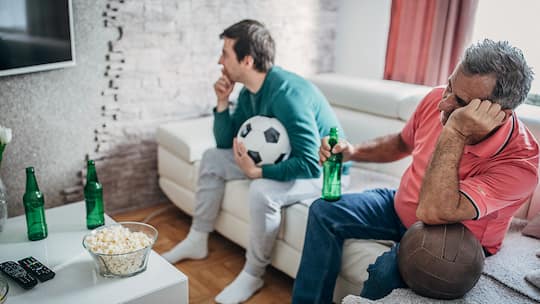 Ein Vater und sein Sohn schauen sich zusammen ein Fussballspiel an, während auf dem Wohnzimmertisch mehrere Flaschen mit Alkohol herumstehen. Dabei sieht der Vater sichtbar benebelt aus.