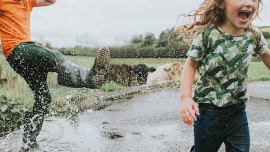 Two cute kids jump in a huge puddle, wearing welly boots. Boy playfully kicks water towards girl as she happily screams. Splashes of water surround the children as they fly towards the camera. Conceptual with space for copy.