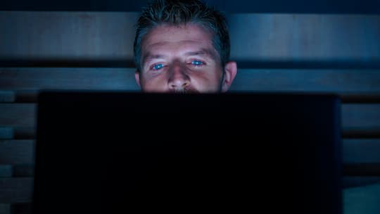 Ein junger Mann liegt im Bett. Sein Gesicht ist erleuchtet vom Bildschirm seines Laptops, der auf seinem Schoss liegt.