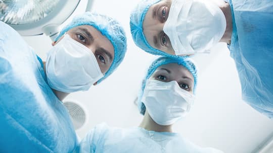 Zwei Chirurgen und eine Chirurgin stehen in einem Operationssaal und blicken von oben auf den vermeintlichen Patienten herab.