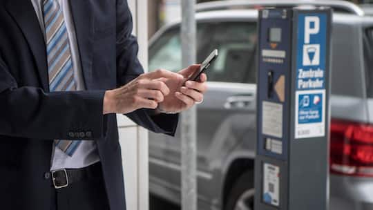 Zu sehen ist ein Mann im Anzug, der gerade etwas auf seinem Handy eintippt. Er befindet sich direkt neben einer Säule für das Bezahlen von Parkgebühren.