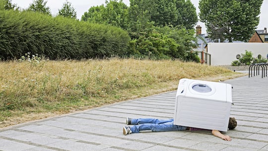 Eine Waschmaschine liegt auf einem Mann auf dem Gehweg - welche Versicherungen es braucht, wenn man in die erste eigene Wohnung einzieht.