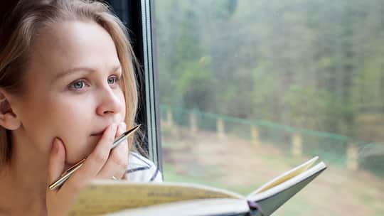 Eine junge Frau hält ein Tagebuch in der Hand, während sie aus dem Zugfenster schaut.
