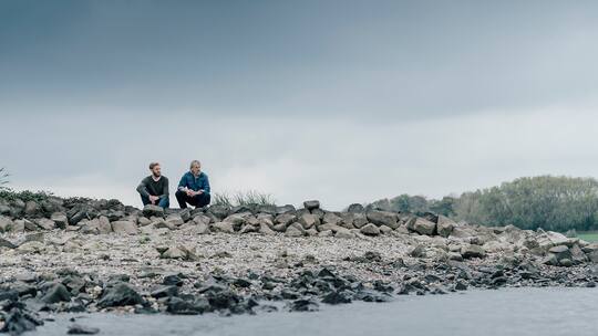 Zwei Männer sitzen an einem Fluss und diskutieren miteinander.