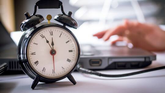 Im Hintergrund arbeitet jemand an einem Laptop. Im Vordergrund ist ein Wecker zu sehen.
