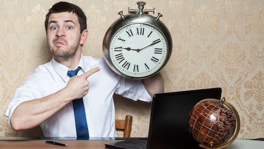 Mann sitzt an einem Schreibtisch und zeigt auf eine Wanduhr, die er in der Hand hält.