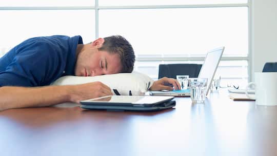 Ein Mann schläft mit einem Kissen als Unterlage auf dem Bürotisch.