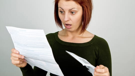 Eine Frau hält in ihrer rechten Hand ein Mahnungsschreiben in der Hand und ist sichtlich über die Nachricht erschrocken.