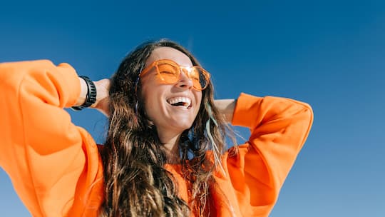 Lachende Frau geniesst die Sonne - Sonnenstrahlung ist gefährlich, aber auch gesund. Die Wissenschaft hat neue positive Effekte entdeckt. Hautärztinnen denken um. Wie man die Sonne dosiert, ohne das Hautkrebsrisiko zu vernachlässigen