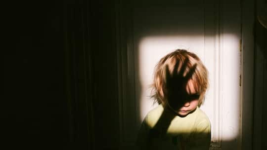 Anonymes kleines Kind, das in einem quadratischen Lichtfleck neben einer Tür in einer alten Wohnung sitzt, mit dem Schatten einer Erwachsenenhand über seinem Gesicht.