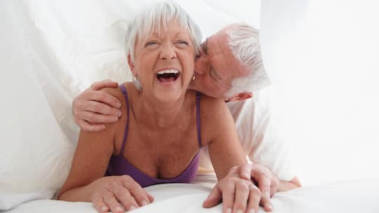 Ein älteres Paar küsst sich unter der Bettdecke und lacht.