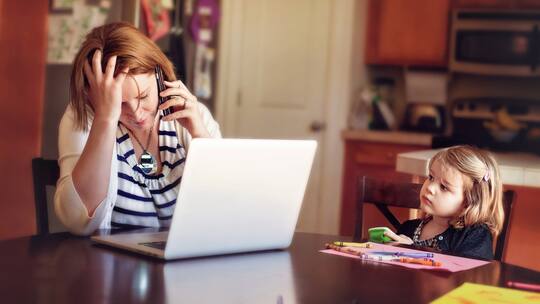 Eine Mutter sitzt am Computer und telefoniert, sie wirkt gestresst. Ihre kleine Tochter sitzt daneben und schaut sie an.