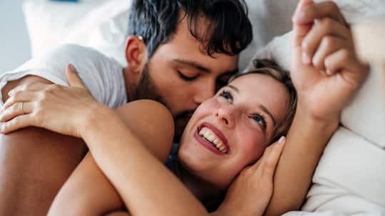 Ein Paar liegt eng umschlungen im Bett, er küsst sie und sie lacht