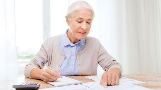 Ältere Frau sitzt an Tisch, sie hält einen Kugelschreiber in der Hand und vor ihr auf dem Tisch liegen Taschenrechner, Schreibblock und Unterlagen.
