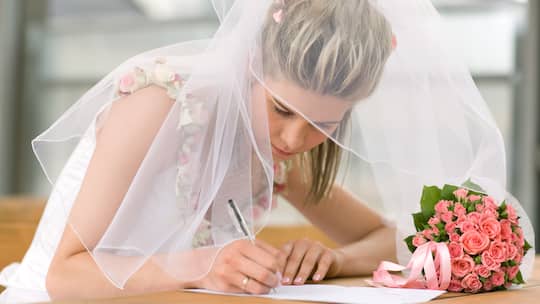 Eine Frau in weissem Brautkleid setzt ihre Unterschrift auf ein Dokument zur Eheschliessung.
