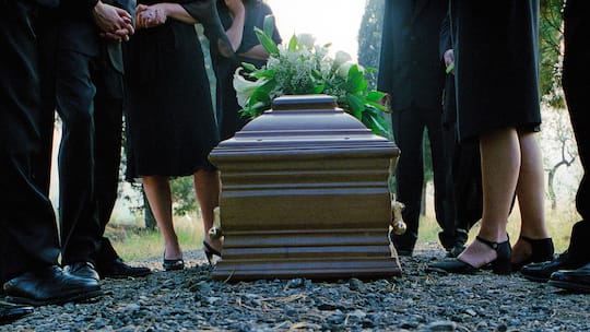 Mit Blumen geschmückter Holzsarg an Trauerfeier und Trauergäste stehen um den Sarg herum.