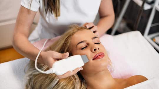 Beautiful young blonde woman receiving professional face ultrasonic peeling treatment. Cosmetology specialist doing modern procedure of cleaning face with ultrasonic scrubber.