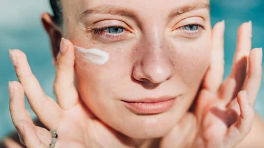A close-up portrait of a woman with her eyes closed and a serene expression, as she applies a small dab of sunscreen or face cream to her cheek.