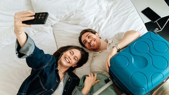 Ein lachendes Paar liegt auf dem Bett im Hotelzimmer und macht ein Selfie.