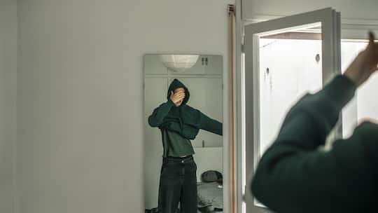 16-year-old young man is dressing up in front of the mirror in his sleeping room. He is a handsome fair-haired boy and he sets his hair so that he is ready to go out. He is wearing a dark green hoodie. He covers his face with his hand.