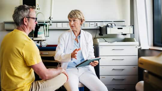 A female doctor in a clinical setting shows a digital X-ray to a patient while engaging in a discussion. The patient, dressed in a yellow shirt, listens attentively, seated across from the doctor.
