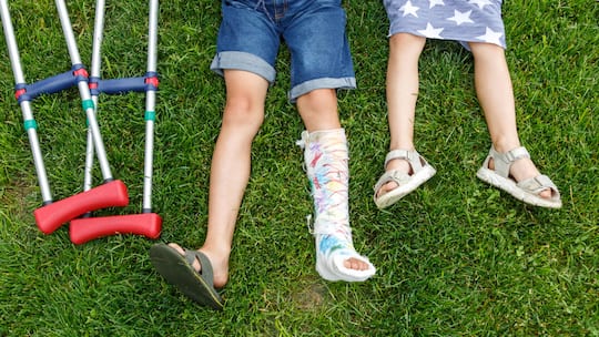 Ein Junge mit gebrochenem Bein im Gips liegt im Gras, neben ihm Krücken.