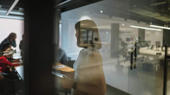 Photo of a young woman using a tablet in her office, preparing for a meeting, and reviewing meeting notes as she is about to start a professional discussion with her coworkers.