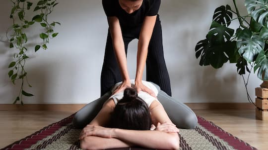 Masseuse giving Thai massage to womans back. Woman in childs pose on futon receiving lumbar massage. Wellbeing concept.