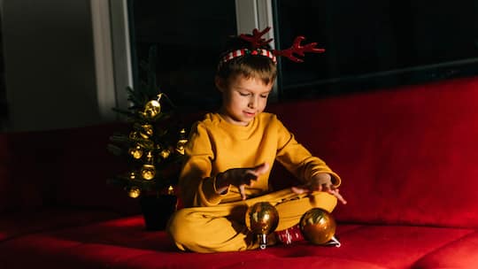 Ein kleiner Junge in einem gelben Pyjama sitzt auf einem roten Sofa neben einem Weihnachtsbaum und packt ein Geschenk aus - Wenn getrennte Paare Weihnachten mit Kindern feiern, stellt sich die Frage, ob sie die Feiertage gemeinsam oder getrennt begehen sollen. Das rät die Beobachter-Expertin für Familienrecht.