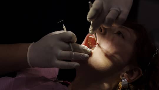 A 47-year-old Caucasian woman receives a dental examination. A dentist's gloved hands use a scaler and a mouth mirror inside her open mouth. The scene is a close-up, low-angle view with dramatic lighting against a black background.