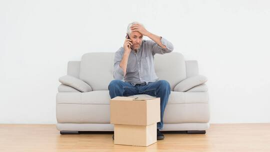 Ein Sofa steht alleine in einer Mietwohnung, davor zwei Umzugskartons. Auf dem Sofa sitzt ein verzweifelter Mann, der sich die Stirn hält und mit dem Handy telefoniert.