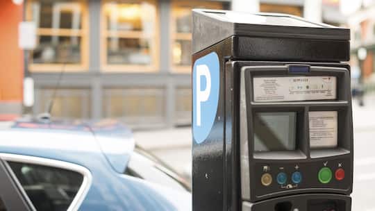 Im Vordergrund sieht man eine Parkuhr und im Hintergrund ist ein parkiertes Auto.