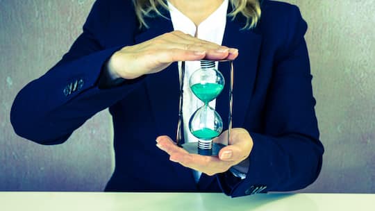 Eine Frau in Business-Kleidung hält vor sich eine Sanduhr in der Hand.
