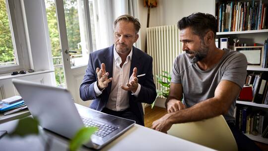 Zwei Männer sitzen vor einem Laptop. Der Mann links mit Anzug benutzt seine Hände, um etwas anschaulich erklären zu können.