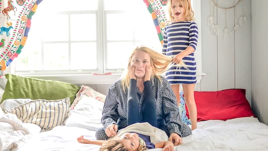 Wie soll man Achtsamkeit in einen ausgefüllten Alltag einbauen? Eine erschöpfte Mutter sitzt mit ihren Kindern auf dem Bett.