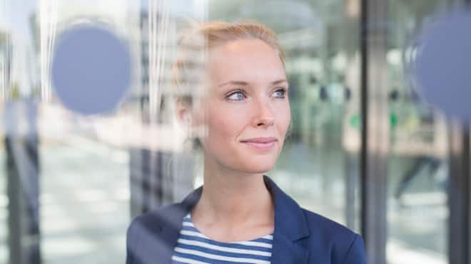 Selber entscheiden Eine Geschäftsfrau mit blauem Blazer schaut zufrieden und selbstbewusst durch ein verglastes Bürogebäude.