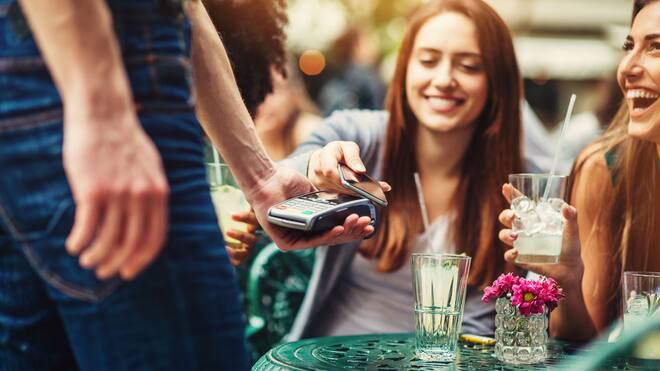 Endlich Geld – und jetzt? Zwei junge Frauen sitzen draussen in einem Café und bezahlen mit dem Handy für ihre Drinks.