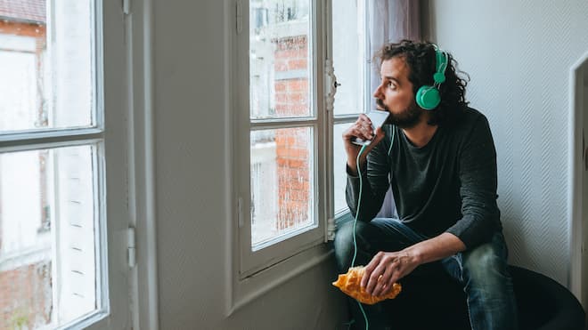 Der Soundtrack bei Liebeskummer Ein Mann sitzt in einer Ecke seiner Wohnung, hört über mit dem Smartphone verbundene Kopfhörer Musik und schaut aus dem verregneten Fenster.