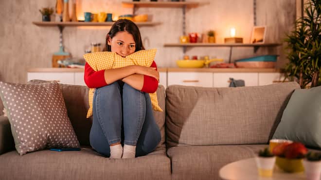 Balsam für das gebrochene Herz Eine junge Frau sitzt traurig und zusammengekauert auf ihrem Sofa und stützt ihren Kopf auf einem Kissen ab.