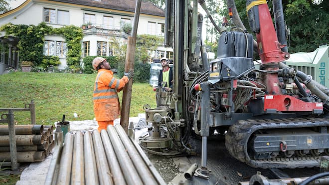 Wie der Wechsel beim Eigenheim gelingt Zwei Mitarbeiter eines Bauunternehmens bohren für eine Wärmepumpe mit Erdsonde in einem Garten eines Hauses.