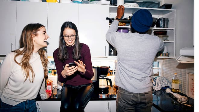Die erste WG – das sollten Sie beachten Eine Wohngemeinschaft von zwei Frauen und einem Mann befinden sich in einer Küche, in der ein kleines Durcheinander herrscht.