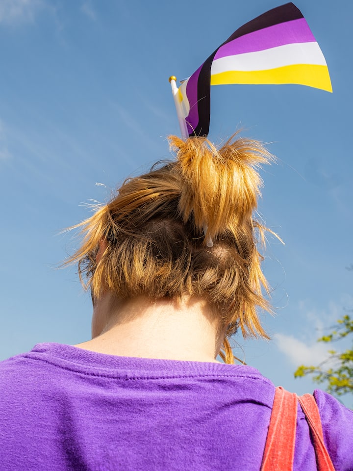 Person mit Trans-Flagge in den Haaren und violettem T-Shirt, von hinten aufgenommen.