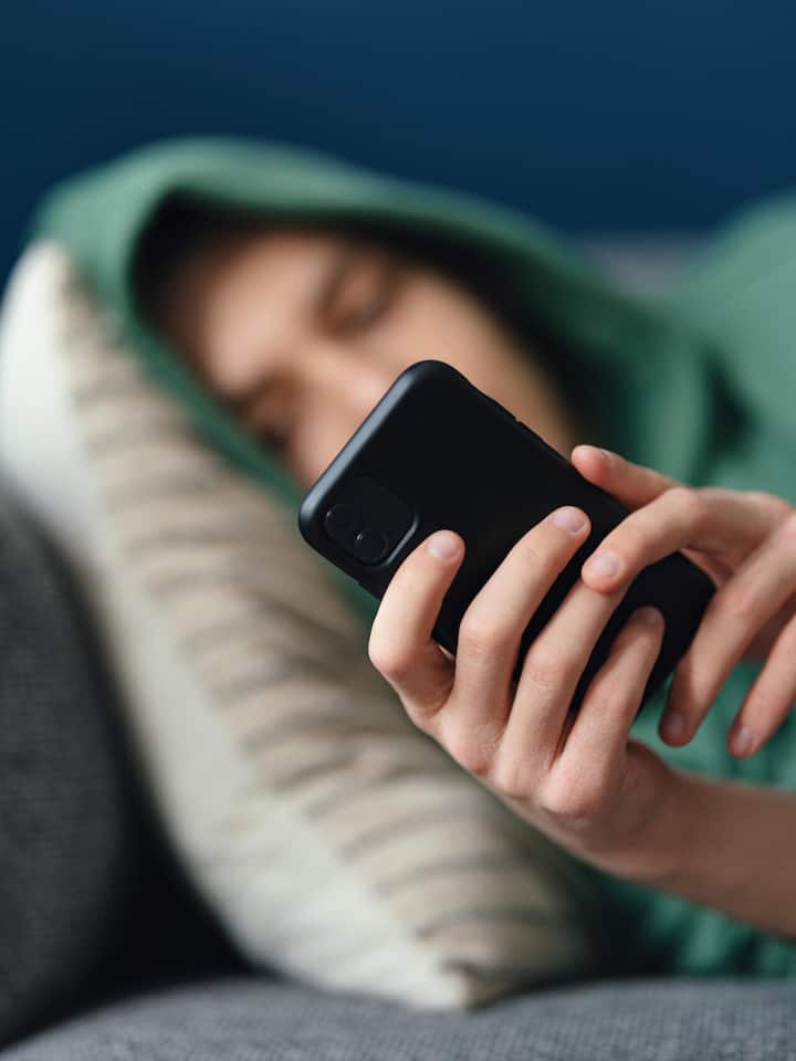 So unterstützen Sie Ihr Kind auf Social Media Ein nicht erkennbarer junger Mann liegt zu Hause seitlich auf dem Sofa und nutzt sein Mobiltelefon zum SMS-Schreiben und Surfen im Internet.
