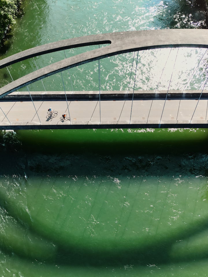 Eine Hängebrücke spannt sich über einem grün glitzernden Fluss, darüber fahren zwei Menschen auf Velos.