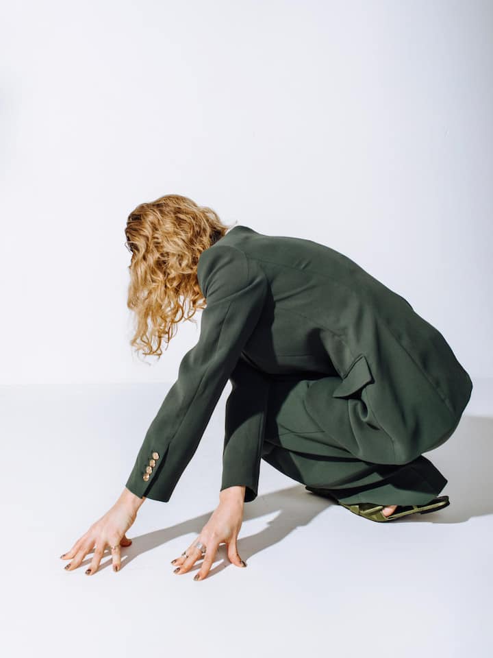 Back view of unrecognizable female fashion model in green suit and pants leaning on floor on white background