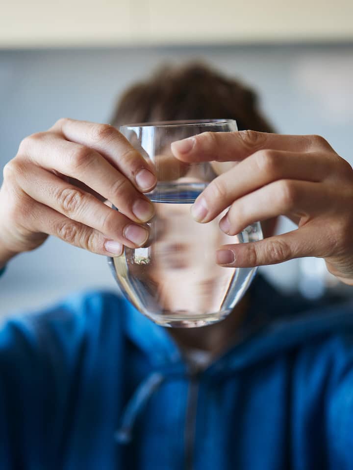 Boy is drinking water from the glass in the kitchen. Focus on glass. Boy is blurred behind.